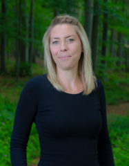 Eine Frau, Anfang 30, mit blondem Haar und dunkelblauem Shirt steht vor Waldbäumen