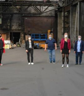 7 Personen stehen in weitem Abstand mit Mundschutz in einer alten Produktionshalle