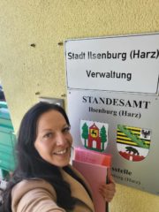 Eine Frau mit dunklem langen Haar steht vor dem Schild der Stadtverwaltung Ilsenburg und hat eine Dokumentenmappe in der Hand und lächelt