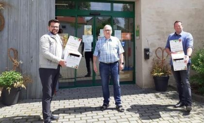3 Männer stehen vor dem Dorfladen und halten etwas in die Kamera