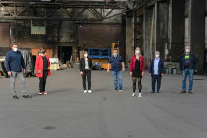 7 Personen stehen in weitem Abstand mit Mundschutz in einer alten Produktionshalle